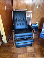 Front view of black tufted upholstered vintage recliner chair with wooden armrests, showing the extendable footrest and some visible wear and mildew.
