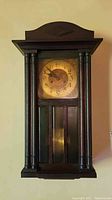 Front view of vintage wooden wall clock showing clock face and pendulum behind glass with detailed wood columns and decorative top
