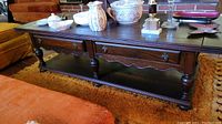 Full front view of the vintage coffee table in room setting, showing top surface with items, drawers, turned legs and lower shelf.