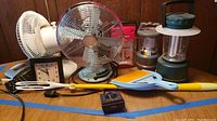 Photo showing two retro tabletop fans, two battery-operated lanterns, desk clocks, adjustable arm lamp, timer, calculator, dustpan with brush on a wood surface