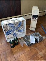 Lot showing box fan with blue blades, DeLonghi heater, wrapped camp stove, and two green propane canisters on wooden floor.