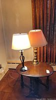 Two table lamps side-by-side on a wooden table. One lamp has a twisted black metal base and white fabric shade, the other has a brass-colored base with pleated beige shade. Both plugged in but untested.