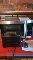 Magic Chef microwave oven MCM990ST stainless steel with digital keypad, along with KitchenSmith black toaster with instruction manual placed on red surface.