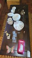 Wide view of all items displayed on wooden surface showing china dishes, candlestick, ashtrays, vase, butterfly ornament, and red ceramic dish