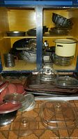 Wide view of cabinet shelf with stacked metal baking pans, Pyrex glass items including a mixing bowl and bread pans, metal kitchen accessories such as graters and sifter, glass and metal lids, and roasting pans. Some metal surfaces show signs of use and patina.
