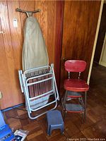 Side view showing ironing board and patio chair folded and leaning against wall near vintage kitchen stool and small step stool on parquet floor.