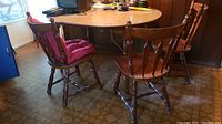 Photo of wooden oval kitchen table with three matching wooden chairs, one chair has a red cushion and damage visible on backrest.