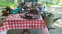 Photo showing various tools, power tools, hand tools, pots, and gardening supplies arranged on a red and white checkered cloth and wooden table outdoors.