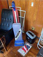 Photo showing the blue ORECK XL vacuum cleaner next to the black and grey paper shredder with plastic bag containing vacuum bags on wooden floor