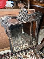 Front view of dark carved wooden mirror frame with detailed ornate top and reflective mirror glass showing patterned carpet reflecting in glass.