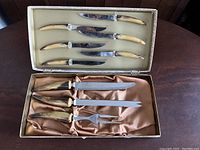 Full view of the carving set in open box showing six knives with yellowish handles in upper section, and carving set pieces in lower section with fabric lining.