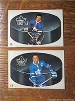 Front of two hockey cards showing Dave Keon and George Armstrong in Toronto Maple Leafs uniforms in front of hockey puck images with player numbers 15 and 13.