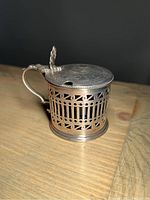 Side view of the sterling silver mustard pot showing its openwork pierced design and handle.