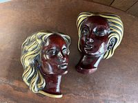Two vintage ceramic female face wall plaques shown from above on a wooden surface. One has wavy hair and the other has striped hair.