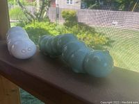 Five ceramic inchworms arranged on a wooden ledge with outdoor greenery visible through a screen. Two cream-colored ones visible in front and one celadon green visible at rear.