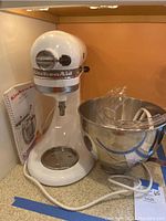 White KitchenAid stand mixer with attached motor housing and speed control lever. Stainless steel mixing bowl placed next to mixer with clear splash guard resting inside the bowl. Instruction manual propped upright behind mixer.