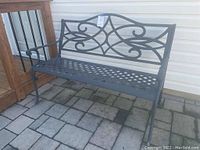 Full view of metal outdoor bench showing lattice seat, armrests, and decorative backrest with scrollwork pattern, placed on stone patio against house wall.