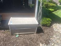 Side view of beige Suncast outdoor storage container in garden mulch beside porch entrance.