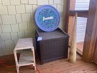 Wide angle photo showing all items - the Suncast storage container, side table, Franklin Flyer disc, and wooden naval artifact on porch wood floor.