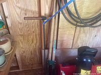 Photo showing UnionTools post hole digger with wooden handles and soil auger leaning against plywood wall with red tool box below, hose coiled on wall