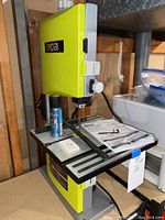 Front view of Ryobi 9 inch band saw. Shows the yellow unit on a workbench with a can of lubricant, hex keys, and operator manual on table surface.