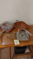 Full view of two Copper Chef fry pans, steamer tray, frying basket and glass lid on wooden surface.