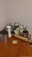 Wide view of baking items: three metal flour sifters, wooden rolling pin, angel food cake pan, two springform pans, three-part white ceramic condiment server
