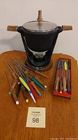 Full assembled cast iron fondue pot on black metal stand with fuel holder, surrounded by various fondue forks with wooden and colored handles, and a box of additional forks.