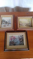 Three framed prints laid flat on a table: two winter rural landscape scenes and one floral print.