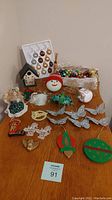 Photo showing various Christmas decorations including colorful glass ornaments in packaging, wooden birdhouse, snowman figure, glittery butterflies, and other hanging ornaments on a wooden table.