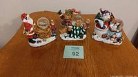 Photo showing three Christmas mini globes on a wooden surface with Santa, bear, and snowman figures and children around each globe.