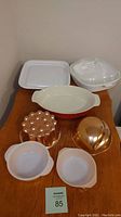 Overall lot on small table showing all items: large oval red Corning Ware casserole, microwave browner grill with lid, small round Corning Ware casseroles, stoneware casserole with glass lid, two copper jelly molds