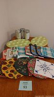 Photo showing 2 pale yellow deviled egg trays, 8 assorted fabric pot holders with various patterns, 3 fabric trivets with floral and sunflower patterns, 1 ceramic tile trivet, 1 braided round trivet, and 3 cylindrical David's Tea cans with metal lids.