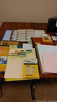 Full assortment of office supplies on a wooden table showing packages of pencils, photo paper, folders, note cards, and a stapler.