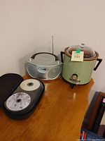 Portable silver radio/CD player with front display and side speakers next to a green Crock-Pot slow cooker with glass lid and black handles, along with cases and boxes of CDs and cassettes on wooden surface.