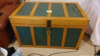 Front and top view of rectangular storage chest featuring natural wood frame and green textured metal paneling, padlocked latch at center front.