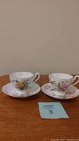 Two Queen Anne fine bone china tea cups and matching saucers with floral designs displayed on wooden surface against beige wall.