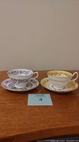 Two Grosvenor tea cups and saucers on a wooden surface, showing side view of cups and saucers, one yellow with gold accents and the other white with pink and green floral designs