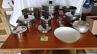 Display of various pots and pans including stainless steel pots with lids, stockpot with pasta insert, frying pans and aluminum kettle on wooden table.