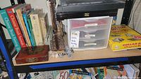 Shelf containing multiple Asian brush painting books, a wooden mahjong box with oriental painting, and a plastic drawer organizer labeled Chinese, Indo, Paintings with assorted art supplies. Also visible is packaging for dollar bill origami kit.
