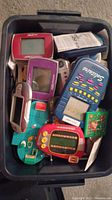 Top view of plastic bin filled with multiple handheld electronic games and accessories in various colors.