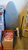 Overview of ironing board, iron, box with fusible interfacing patterns and other sewing materials on floor by plastic drawers