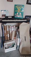 Photo showing the Lowrey V-60 electronic keyboard on a metal stand with books displayed upright behind it plus a protective cover and rolls of material stored underneath the stand.