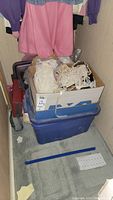Photo showing the cardboard box containing lace, fabrics and other textile items on top of a blue storage bin, with some hanging clothes partially visible above.