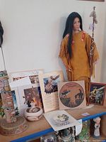Photo showing entire lot including the porcelain doll in traditional dress, the totem pole, painted pottery, and multiple Native American themed prints and artwork.