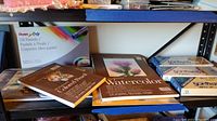 Photo of art supplies on shelf showing a box of Pentel Arts oil pastels, watercolor sketchpad, colored pencil sketchpads, and watercolor pencil pads.