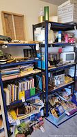 Two metal shelving units shown in a cluttered room with various books, papers, small electronics and tools on shelves. Units are black metal with multiple shelves. Contents on shelves not included.