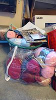 Wide view of assorted yarn balls in plastic bags with patterns and crocheting tools in green basket and bag.