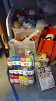 Multiple color yarn skeins stacked on floor with plastic storage bins and orange bags in background