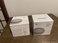 Two boxes of Google Home Mini smart speakers side-by-side on a table; one box is unopened and sealed, the other opened showing the speaker inside.